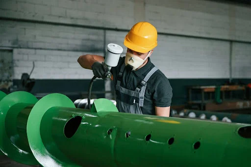 Industrial worker applying protective coating to metal equipment in a manufacturing facility to prevent corrosion and extend lifespan.